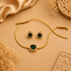 A gold-plated necklace and matching earring set featuring vibrant green emerald stones encircled by sparkling white crystals, displayed on a beige surface with satin and floral accents. Available in the UK at theindianshelf.co.uk.