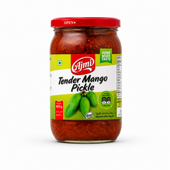 A professional studio product shot of a clear glass jar containing Ajmi Tender Mango Pickle, standing vertically against a solid white background The jar is filled with a rich, deep red spiced pickle mixture and topped with a glossy red metal lid printed with an "OPEN" arrow indicator. The light green and white wrap-around label features the red oval "Ajmi" logo above the text "Tender Mango Pickle" written in a stylized black font.This product is available in the UK at theindianshelf.co.uk.