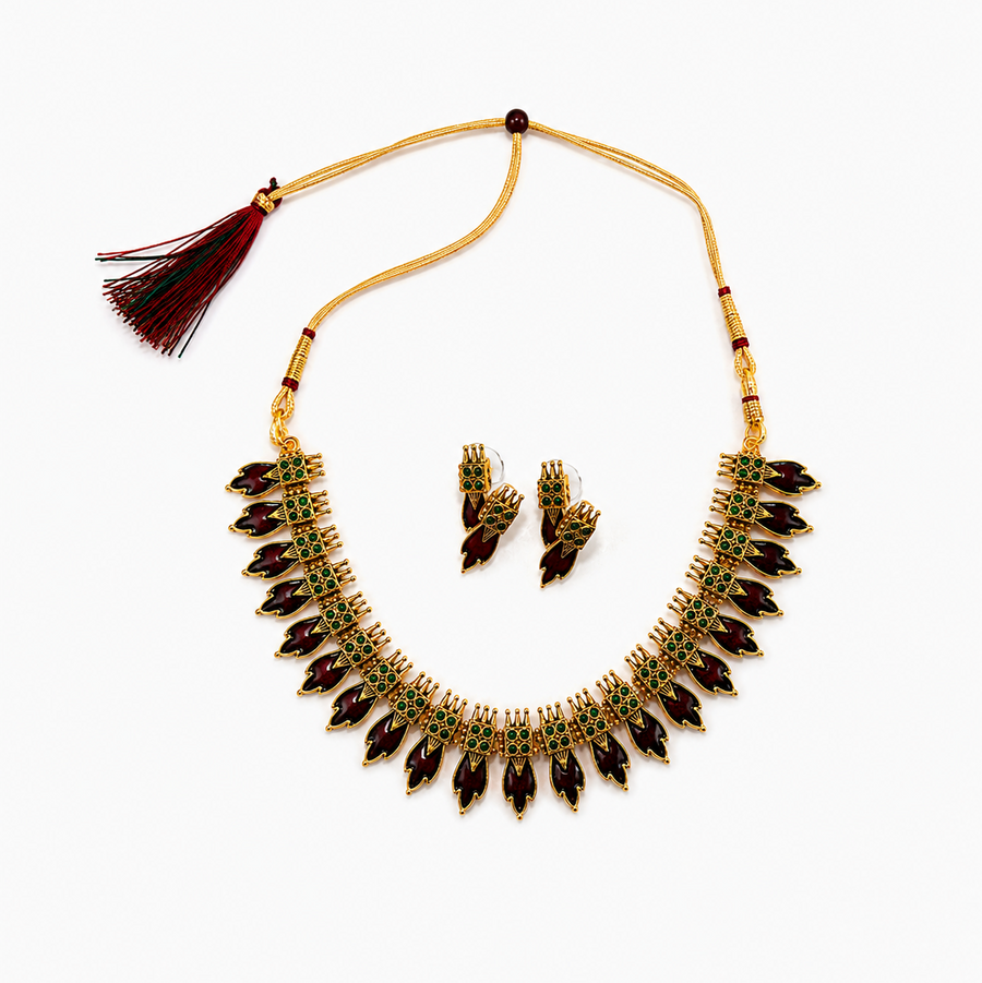 An antique gold-plated temple jewelry set featuring a leaf-motif necklace and matching earrings adorned with deep maroon and emerald green kemp stones, centered on a solid white background. Available in the UK at theindianshelf.co.uk.