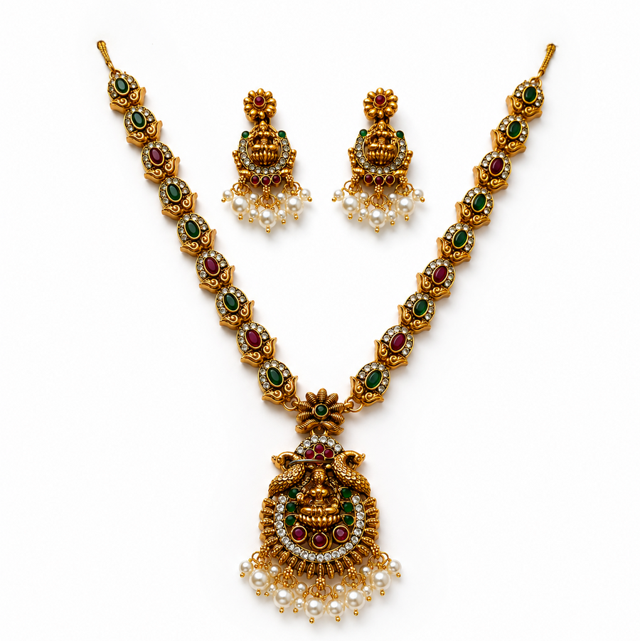 A studio product shot of an antique gold-plated Temple Jewelry Set isolated on a white background. The long necklace and matching earrings feature a Goddess Laxmi pendant decorated with red, green, and white stones and finished with dangling pearls. Available in the UK at theindianshelf.co.uk.