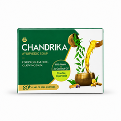 Studio product shot of a rectangular box of Chandrika Ayurvedic Soap against a solid white background. The packaging features a dual-toned design split diagonally, with dark green on the left and crisp white on the right. The bold white brand name "CHANDRIKA" is printed on the green side directly above the subtitle "FOR PROBLEM-FREE, GLOWING SKIN.". A vibrant yellow circular badge reads "With Neem & 2x Coconut Oil Double Ayurveda This product is available in the UK at theindianshelf.co.uk.