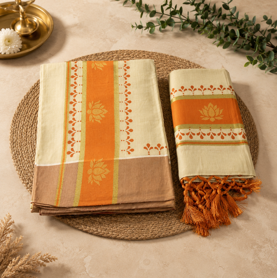 A studio flatlay shot of a folded cream Kerala Dhavani Set arranged on a round jute mat. The cotton garments feature a bright orange border design accented with golden lotus prints and fine orange fringe tassels. A traditional brass lamp and a green leaf stem decorate the background. Available in the UK at theindianshelf.co.uk.