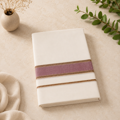 A studio product shot of a neatly folded cream Kerala Double Mundu placed diagonally on a beige tiled surface. The fabric features a bold horizontal border consisting of a wide mauve-purple band framed by thin gold and maroon stripes. A small textured ceramic vase and green leaves frame the layout. Available in the UK at theindianshelf.co.uk.