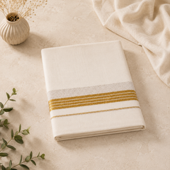 A studio product shot of a neatly folded cream Kerala Double Mundu placed diagonally on a beige marble surface. The cotton fabric displays a horizontal border pattern composed of rich golden stripes running parallel to a broad silver woven band. A small fluted ceramic vase and a soft cream fabric drape frame the top layout. Available in the UK at theindianshelf.co.uk.