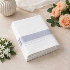 A studio product shot of a neatly folded white Kerala Double Mundu placed diagonally on a beige tiled surface. The fabric features a horizontal border pattern made of light silver-grey stripes. Peach roses decorate the upper right corner, and white jasmine flowers sit on the lower left. Available in the UK at theindianshelf.co.uk.