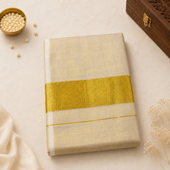A studio flatlay shot of a folded shimmering golden-beige Kerala Gold Tissue Saree on a light marble surface. The fabric features a metallic sheen, accented by a central wide gold kasavu band and a thin gold pinstripe. A brass dish with white beads and a wooden box rest in the background. Available in the UK at theindianshelf.co.uk.