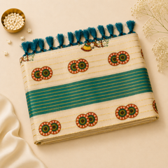 A studio flatlay shot of a folded cream-gold Kerala Tissue Saree on a light beige backdrop. The metallic gold body features a wide teal-blue center stripe, circular ethnic geometric motifs, a traditional peacock design, and rich teal fringe tassels. Available in the UK at theindianshelf.co.uk.