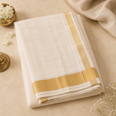 A studio flatlay shot of a neatly folded cream-white Kerala Kasavu Saree arranged on a warm beige surface. The cotton fabric features a broad, brilliant horizontal and vertical golden zari border. The top layout is elegantly framed by a small brass bowl of white jasmine blossoms and a soft cream textile drape. Available in the UK at theindianshelf.co.uk.