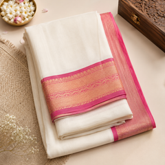 A studio flatlay shot of a neatly folded cream Kerala Kasavu Saree on a light beige background. The cotton fabric features a bright pink border decorated with woven rose-gold zari floral patterns. The layout includes a carved wooden box, a brass bowl of jasmine blossoms, and a fringed placemat. Available in the UK at theindianshelf.co.uk.