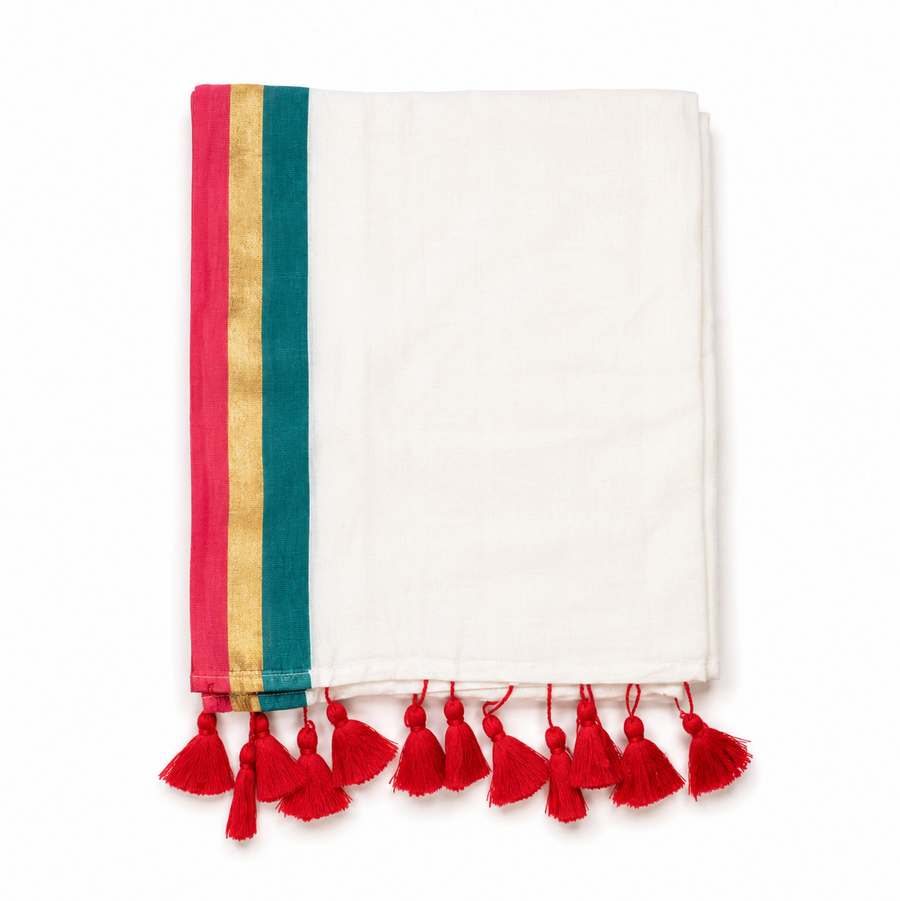 A neatly folded off-white mulcotton saree centered on a solid white background, featuring a tri-color vertical border with magenta pink, golden kasavu, and teal green stripes, finished with bright red handcrafted tassels along the bottom fringe. Available in the UK at theindianshelf.co.uk.