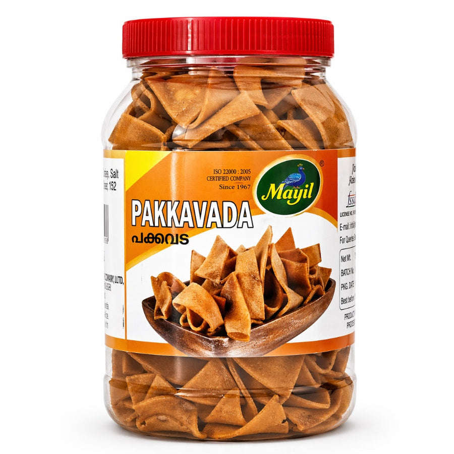 A vertical studio shot of a large, clear plastic jar filled with Mayil Pakkavada. The jar features a bright red screw-top lid and a vibrant orange and yellow label. The label prominently displays the "Mayil" logo—a green oval featuring a blue peacock. An illustration shows a rustic wooden bowl filled with the golden-brown, ribbon-like snacks. The jar is set against a clean, plain white background. This product is available in the UK at theindianshelf.co.uk.