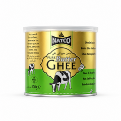 A studio product shot of a cylindrical 500g tin of Natco Pure Butter Ghee standing vertically against a solid white background The tin features a half-metallic-gold and half-bright-green split color scheme, capped by a semi-translucent plastic lid. The top center displays the black "NATCO" corporate shield logo. Below it, inside a golden ornate frame, the text reads "PURE Butter GHEE" alongside its Arabic translation. This product is available in the UK at theindianshelf.co.uk.