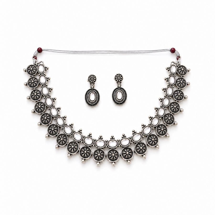 A studio product shot of an antique oxidized silver Choker Necklace and Earrings Set isolated on a white background. The jewelry features sculpted floral metalwork details set with clear white stones. Available in the UK at theindianshelf.co.uk.