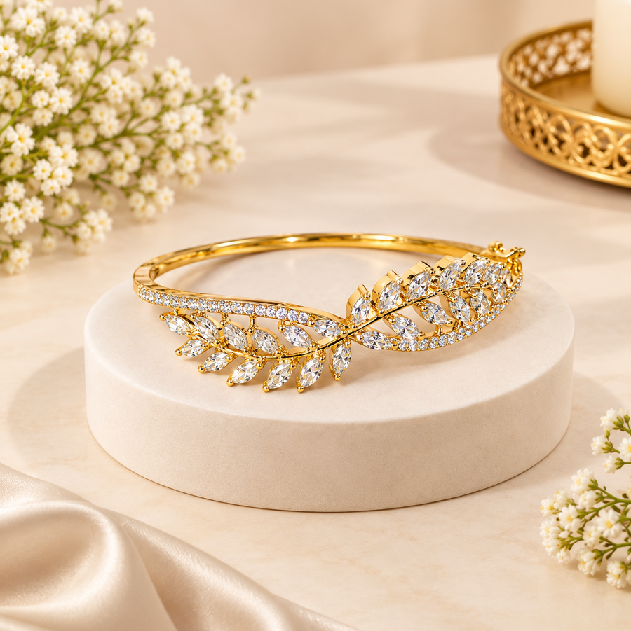 A polished gold openable bangle featuring an elaborate leaf and vine design encrusted with sparkling white cubic zirconia stones, displayed on a neutral platform. Available in the UK at theindianshelf.co.uk.
