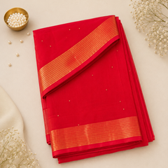 A studio flatlay shot of a neatly folded crimson red Kerala Cotton Saree with subtle gold dots. The fabric features a prominent horizontal border with fine parallel gold zari stripes. A gold dish with white pearls and dried flowers decorate the background. Available in the UK at theindianshelf.co.uk.