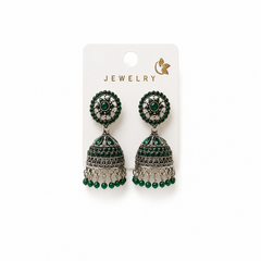 A studio product shot of a pair of antique silver oxidized Jhumka Earrings pinned to a white card against a white background from image_256199.jpg. The drop earrings feature a detailed dome bell accented with green stones and beads. Available in the UK at theindianshelf.co.uk.