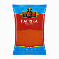 A 100g pouch of TRS Paprika centered on a solid white background, featuring a bright red and blue packaging design with the TRS logo and a clear window displaying finely ground red paprika powder. This product is available in the UK at theindianshelf.co.uk.