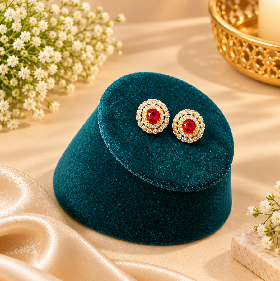 Gold-plated circular halo stud earrings centered with a ruby-red stone and surrounded by white CZ crystals on a teal velvet stand. Available in the UK at theindianshelf.co.uk.