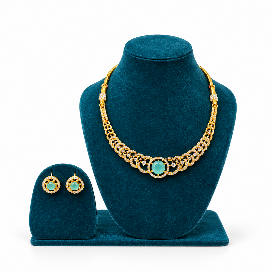 A gold-plated necklace set with a central mint-green stone and sparkling CZ links, displayed on a peacock-blue velvet bust with matching studs. Available in the UK at theindianshelf.co.uk.
