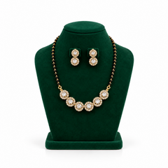 A gold-plated mangalsutra necklace with black beads and a row of circular CZ stone medallions displayed on a green velvet bust with matching earrings. Available in the UK at theindianshelf.co.uk.
