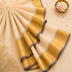 A studio flatlay shot of a folded golden-beige Kerala Tissue Saree on a light cream surface. The fabric displays a metallic sheen, framed by a wide gold kasavu border with dark charcoal grey edges. Available in the UK at theindianshelf.co.uk.