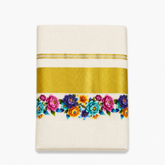 A close-up folded view of an off-white Kerala cotton kasavu set mundu centered on a solid white background, displaying a horizontal metallic gold zari band alongside a vibrant floral print border in pink, purple, turquoise, and yellow. Available in the UK at theindianshelf.co.uk.