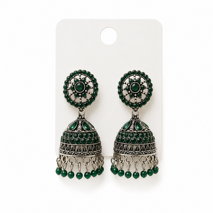 A studio product shot of a pair of antique silver oxidized Jhumka Earrings pinned to a white card against a white background from image_256199.jpg. The drop earrings feature a detailed dome bell accented with green stones and beads. Available in the UK at theindianshelf.co.uk.