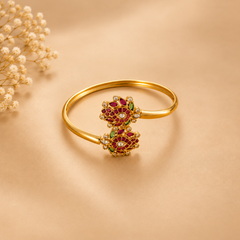 A polished gold-plated ethnic bangle featuring an intricate dual peacock motif adorned with ruby-red and green stones, styled on a warm beige background. Available in the UK at theindianshelf.co.uk.