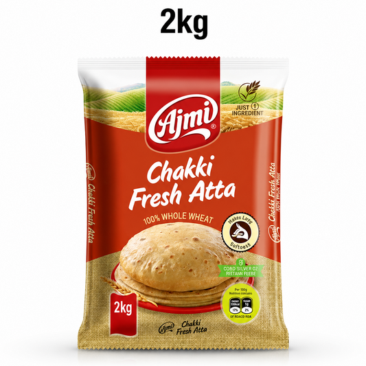 A professional studio product shot of a 2kg pillow pouch of Ajmi Chakki Fresh Atta standing vertically against a solid white background, with a "2kg" text label floating centered above it. The upper portion of the package shows a lush illustration of golden wheat fields beneath a clear sky, segmented by a central red stripe housing the white "Ajmi" logo. This product is available in the UK at theindianshelf.co.uk.