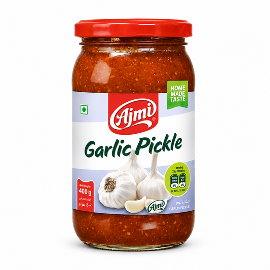 A professional studio shot of a 400 g glass jar of Ajmi Garlic Pickle. The jar is filled with a thick, textured, deep reddish-brown pickle containing visible spices. The label features a clean white and light purple design with the bold red "Ajmi" logo at the center. The label includes a green "HOME MADE TASTE" seal, nutritional information bubbles, and the green vegetarian symbol. This product is available in the UK at theindianshelf.co.uk.