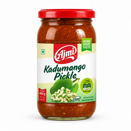 A professional studio shot of a 400 g glass jar of Ajmi Kadumango Pickle. The jar is filled with a rich, dark reddish-brown textured pickle. The label features a light cream and green background with the red "Ajmi" logo at the top center. Below the branding, an illustration shows a whole green mango next to a pile of small, diced green mango pieces. A "HOME MADE TASTE" banner is visible on the right, along with nutritional information bubbles. This product is available in the UK at theindianshelf.co.uk.