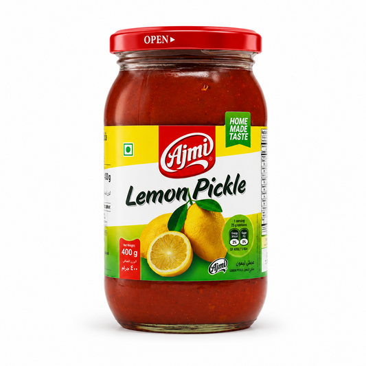 A professional studio shot of a 400 g glass jar of Ajmi Lemon Pickle. The jar is filled with a bright, thick reddish-orange pickle. The label features a cheerful yellow and green design with the red "Ajmi" logo at the center. Below the brand name, a graphic shows two whole yellow lemons and one sliced half with fresh green leaves. The label includes a "HOME MADE TASTE" seal, the green vegetarian symbol, and nutritional information bubbles. This product is available in the UK at theindianshelf.co.uk.