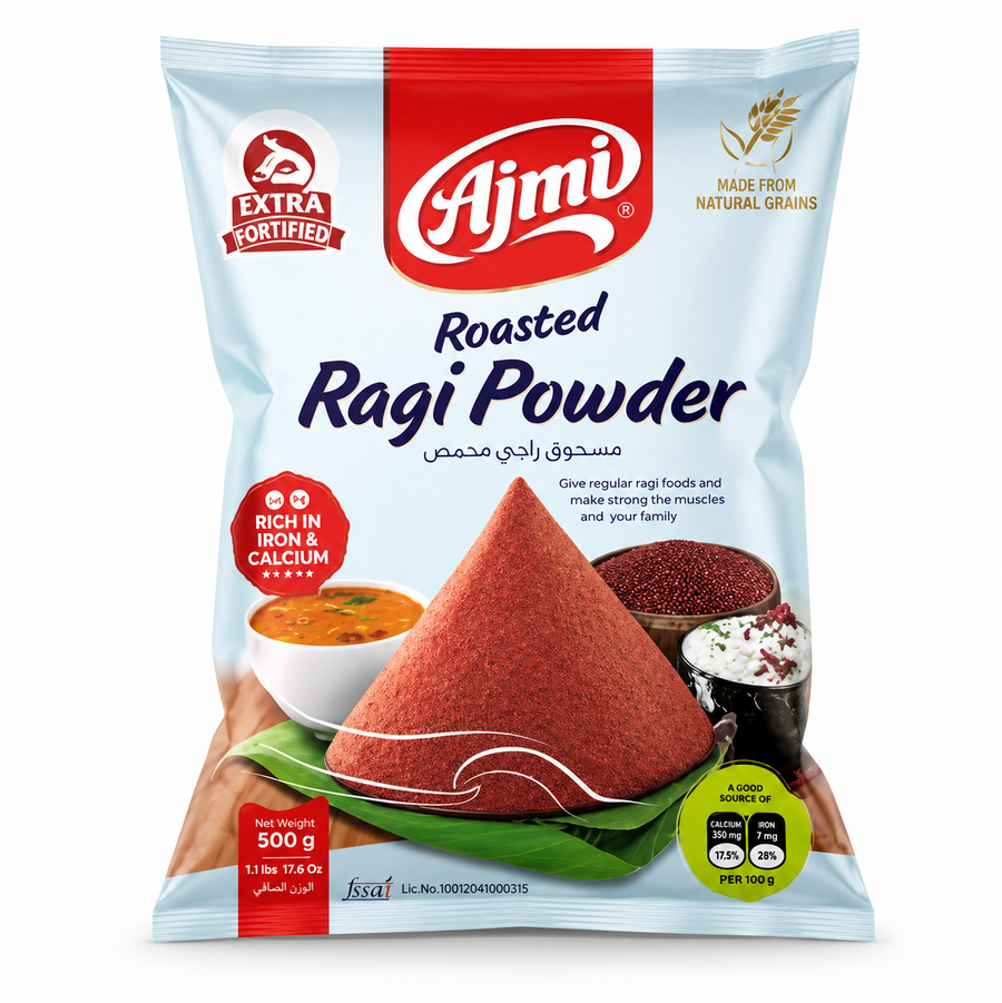 A professional studio shot of a 500 g bag of Ajmi Roasted Ragi Powder as shown in image_d3d078.jpg. The packaging is a light sky-blue with a prominent red top section containing the "Ajmi" logo. The center of the bag features a large, conical mound of deep-red roasted ragi powder resting on a green banana leaf. Various dishes like ragi porridge and grains are pictured in the background. 