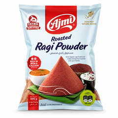 A professional studio shot of a 500 g bag of Ajmi Roasted Ragi Powder as shown in image_d3d078.jpg. The packaging is a light sky-blue with a prominent red top section containing the "Ajmi" logo. The center of the bag features a large, conical mound of deep-red roasted ragi powder resting on a green banana leaf. Various dishes like ragi porridge and grains are pictured in the background. 