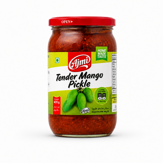 A professional studio product shot of a clear glass jar containing Ajmi Tender Mango Pickle, standing vertically against a solid white background The jar is filled with a rich, deep red spiced pickle mixture and topped with a glossy red metal lid printed with an "OPEN" arrow indicator. The light green and white wrap-around label features the red oval "Ajmi" logo above the text "Tender Mango Pickle" written in a stylized black font.This product is available in the UK at theindianshelf.co.uk.