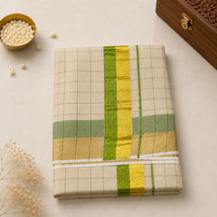 A flatlay product shot of a neatly folded golden-beige Kerala Gold Tissue Saree featuring a green check pattern. The fabric displays a metallic sheen, accented by a central emerald green border with a shimmering gold zari band. A carved wooden box and a brass dish with jasmine flowers rest in the background. Available in the UK at theindianshelf.co.uk.
