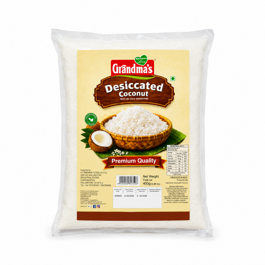 A professional studio product shot of a 400g clear pillow pouch of Grandma's Premium Quality Desiccated Coconut isolated against a pure white background in image_7cc1e5.jpg. The transparent packaging showcases the fine, snowy white desiccated coconut inside, covered by a large light-yellow and brown label displaying a woven bowl overflowing with shredded coconut next to a halved fresh coconut. This product is available in the UK at theindianshelf.co.uk.