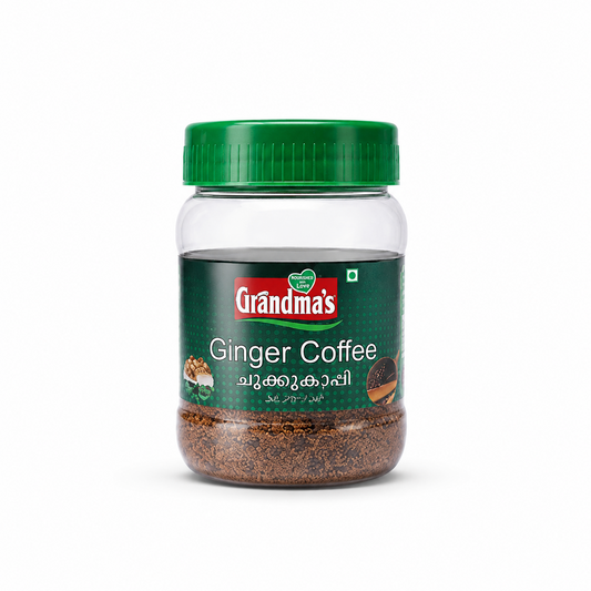 A professional studio shot of a transparent plastic jar of Grandma's Ginger Coffee The jar is partially filled with a coarse, dark-brown coffee and spice blend, and it is sealed with a green ribbed plastic lid. The label features a dark forest-green textured background with the red and white "Grandma's" logo centered near the top. The product title "Ginger Coffee" is printed in clean white text alongside its Malayalam name. This product is available in the UK at theindianshelf.co.uk.