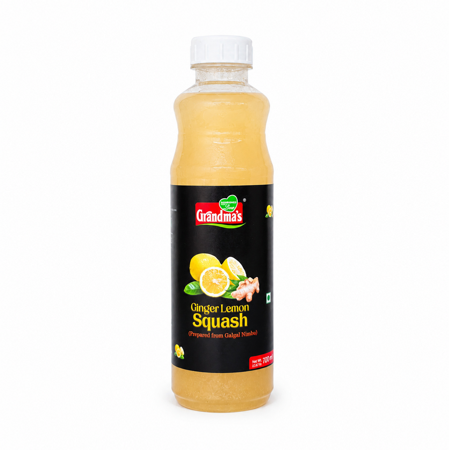 A professional studio product shot of a 700ml bottle of Grandma's Ginger Lemon Squash isolated against a pure white background in image_7afc25.jpg. The transparent bottle reveals a light golden-yellow liquid inside and features a bold black label decorated with graphics of whole and halved fresh lemons, a piece of ginger root, and the red and green brand logo. This product is available in the UK at theindianshelf.co.uk.