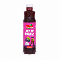 A professional studio product shot of a 700ml bottle of Grandma's Grape Squash isolated against a pure white background in image_7c6042.jpg. The transparent plastic bottle reveals a deep purple-red liquid inside and features a bright pink label decorated with illustrations of fresh dark grapes, a farmer carrying a fruit basket, and the red and green brand logo. This product is available in the UK at theindianshelf.co.uk.