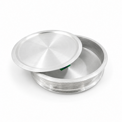 A professional studio product shot of a silver-colored Aluminium Uruli with its matching flat lid partially resting across the top, isolated against a pure white background in image_2319e2.jpg. The shallow, wide-mouthed traditional cooking pot features a clean brushed metallic finish and sturdy construction, displayed under balanced studio lighting for high-end digital listings. This product is available in the UK at theindianshelf.co.uk.