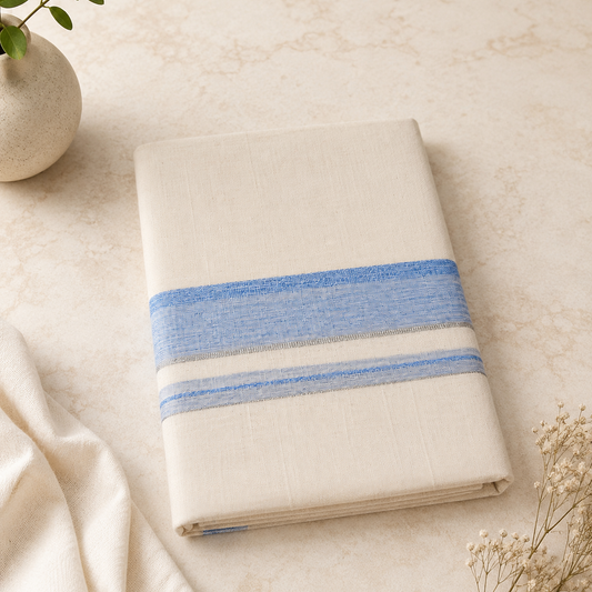 A studio product shot of a neatly folded cream Kerala Double Mundu placed diagonally on a beige marble surface. The cotton fabric features a horizontal border pattern consisting of a textured light blue band flanked by thin silver stripes. A soft white textile drape and delicate dried flowers sit in the background. Available in the UK at theindianshelf.co.uk.