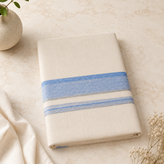A studio product shot of a neatly folded cream Kerala Double Mundu placed diagonally on a beige marble surface. The cotton fabric features a horizontal border pattern consisting of a textured light blue band flanked by thin silver stripes. A soft white textile drape and delicate dried flowers sit in the background. Available in the UK at theindianshelf.co.uk.