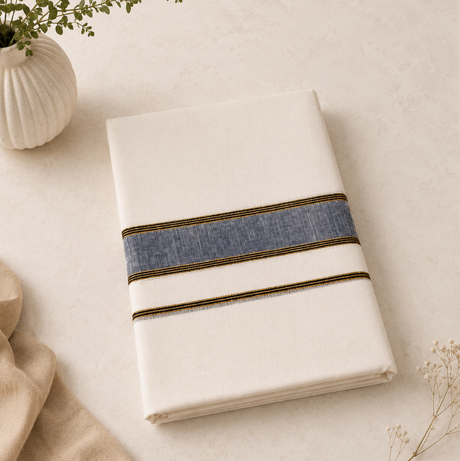 A studio product shot of a neatly folded cream Kerala Double Mundu placed diagonally on a light textured surface. The cotton fabric features a horizontal border pattern consisting of a textured indigo blue band flanked by thin golden stripes. A white fluted vase with a green sprig decorates the top left corner. Available in the UK at theindianshelf.co.uk.