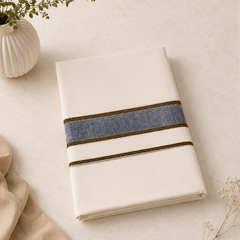 A studio product shot of a neatly folded cream Kerala Double Mundu placed diagonally on a light textured surface. The cotton fabric features a horizontal border pattern consisting of a textured indigo blue band flanked by thin golden stripes. A white fluted vase with a green sprig decorates the top left corner. Available in the UK at theindianshelf.co.uk.