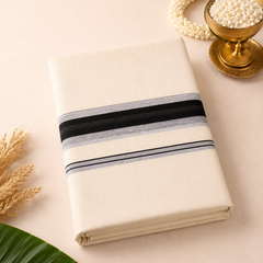 A studio product shot of a neatly folded Kerala Double Mundu styled on a light beige surface. The off-white cotton fabric features a bold horizontal border made up of thick black and grey woven bands. The arrangement is decorated with a brass bowl of jasmine flowers and a green banana leaf corner. Available in the UK at theindianshelf.co.uk.