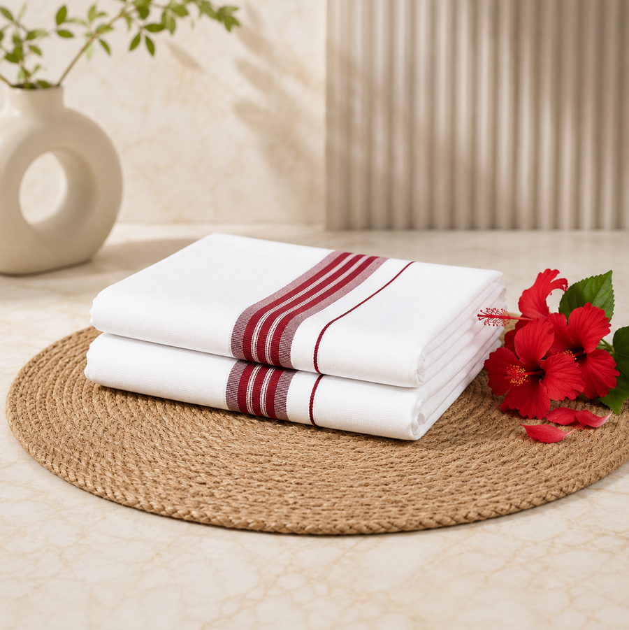 A studio product shot of a neatly folded white Kerala Double Mundu stacked on a round jute placemat. The fabric features a clean horizontal border pattern made of deep red-maroon stripes. Bright red hibiscus flowers sit on the right side of the arrangement. Available in the UK at theindianshelf.co.uk.