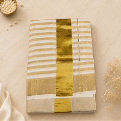 A studio flatlay shot of a folded shimmering golden-beige Kerala Gold Tissue Saree on a light marble surface. The fabric features a metallic sheen, accented by a central wide gold kasavu band and a thin gold pinstripe. A brass dish with white beads and a wooden box rest in the background. Available in the UK at theindianshelf.co.uk.