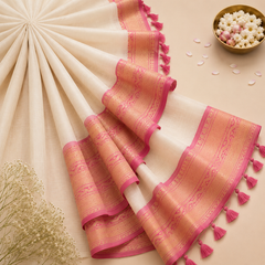 A flatlay product shot of a cream-gold Kerala Kuthampully Saree fanned out diagonally on a beige backdrop. The textile features a magenta-pink border woven with golden zari patterns, green pinstripes, and hanging fringe tassels. A brass bowl with jasmine and purple flowers sits in the upper right corner. Available in the UK at theindianshelf.co.uk.