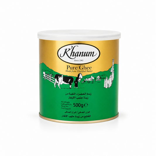 A 500g tin can of Khanum Pure Ghee centered on a solid white background, featuring a gold and green packaging design with the Khanum logo and an illustration of a cow and a traditional figure in a village setting. This product is available in the UK at theindianshelf.co.uk.